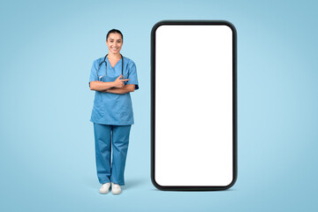 Confident female nurse in blue uniform with stethoscope beside blank phone screen, mockup