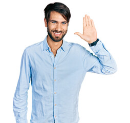 Hispanic man with blue eyes wearing business shirt waiving saying hello happy and smiling, friendly welcome gesture