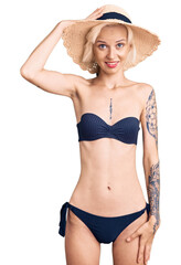 Young blonde woman with tattoo wearing bikini and summer hat confuse and wonder about question. uncertain with doubt, thinking with hand on head. pensive concept.