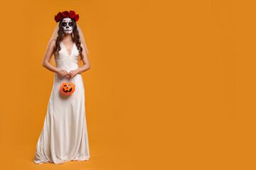 Young woman in scary bride costume with sugar skull makeup, flower crown and pumpkin bucket on orange background, space for text. Halloween celebration