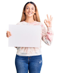 Beautiful young woman holding blank empty banner doing ok sign with fingers, smiling friendly gesturing excellent symbol