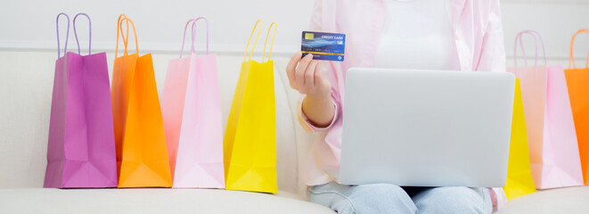 Young asian woman sitting on sofa using laptop computer shopping online with credit card while...
