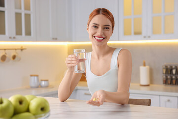 Happy young woman with glass of water and pills at table in kitchen. Weight loss