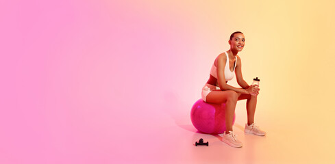A smiling woman with a shaved head takes a break on a fitness ball, holding a water bottle