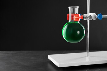Retort stand and laboratory flask with liquid on table against black background, closeup. Space for text