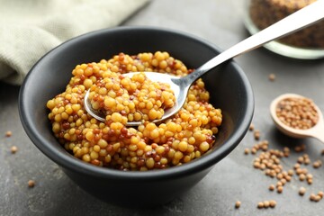 Whole grain mustard, spoon and dry seeds on grey table, closeup