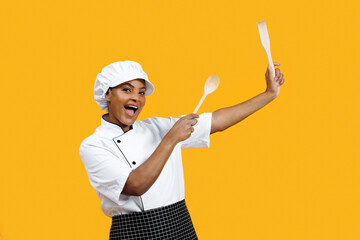 Cheerful young black female chef holding wooden spoons like drumsticks