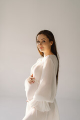 Cute young Pregnant girl sitting on the white studio in warm light with hands on pregnant stomach on wall background with shadow. Pregnant concept, free space, enjoying pregnancy. Pregnant body.