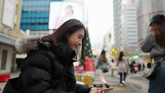 韓国のソウルの明洞の寒い冬に座ってスマートフォンを操作するロングヘアーの30代の韓国人男性のスローモーション映像 Slow-motion video of a long-haired Korean man in his 30s operating a smartphone in the cold winter of Myeong-dong, Seoul, South Korea