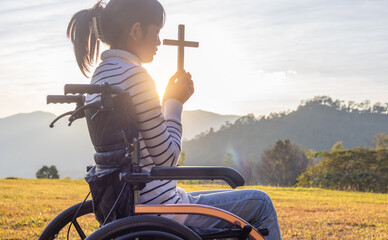 Christian, Christianity, Religion copy space background. Silhouette girl in wheelchair praying and holding christian cross for worshipping God at sunset background.