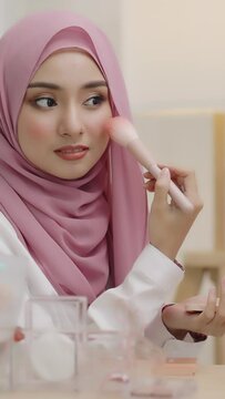 Vertiocal footage Young asian beautiful muslim woman with hijab looking mirror applying makeup with brusher of cheek daily routine at home.