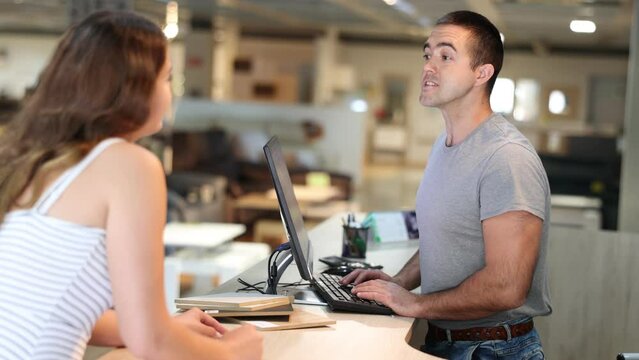 Focused young salesman of furniture showroom working with computer at information desk, advising female buyer on assortment . High quality 4k footage