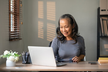 Senior Asian woman happily working on her laptop at home in the living room.