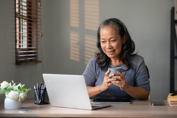 Senior asian woman holding coffee using laptop computer, working, chatting, social media internet at home.