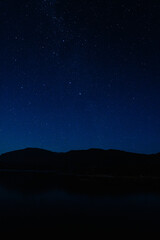 Dark blue starry sky, mountain silhouette, and tranquil lake at night.