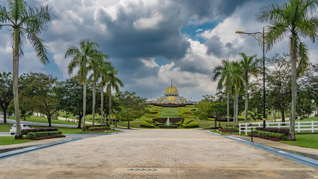 Tall royal palm trees grow in a row on the sides of the paved road. There is a green lawn ahead, decorative flower beds. There is a royal palace Istana Negara on the hill.  Domes with spires 