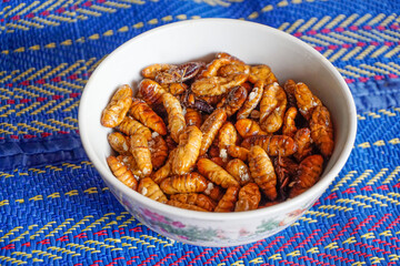 Lots of crispy fried chrysalis in a white bowl placed on a mat. chrysalis are a food that is very high in protein.