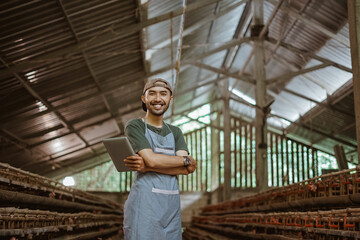 smiling Asian male worker with hands crossed holding a tablet on a chicken farm