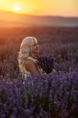 Fototapeta na wymiar Blonde woman poses in lavender field at sunset. Happy woman in white dress holds lavender bouquet. Aromatherapy concept, lavender oil, photo session in lavender