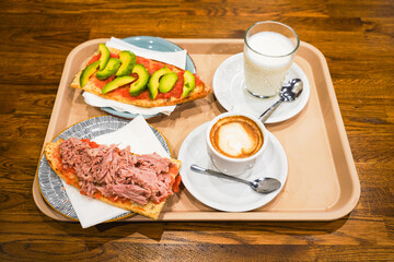 Fresh avocado and tuna toasts with coffee and milk on a cafeteria tray.