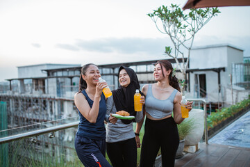 three Asian girls joking around in workout clothes while drinking and snacking after an outdoor...