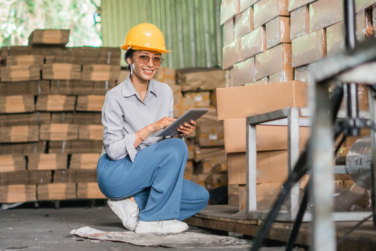 smiling female factory worker wearing safety helmet while squatting holding a tablet in warehouse