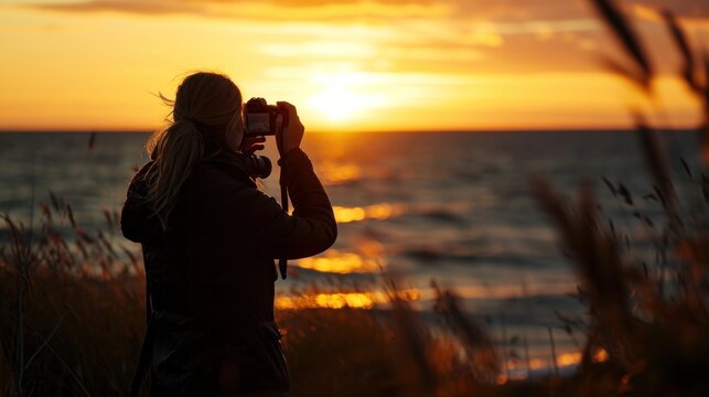  a woman taking a picture of the sun setting over the ocean with a camera in front of her and a body of water in the background.