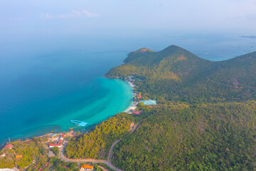 Fototapeta na wymiar Tropical island with a sandy azure bay and pier and a beach village settlement surrounded with roads by exotic green forests trees hills, aerial view