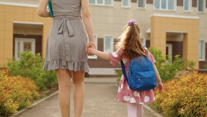 Little girl pupil with backpack going to elementary school first lesson with mother holding hands back view closeup. Cute female kid child and woman mom walking autumn campus schoolyard to classroom