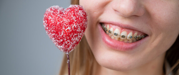 Cute woman with braces on her teeth holds a candy in the form of a heart on white background....