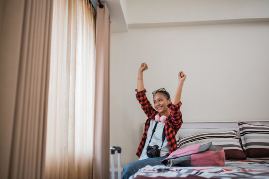 happy female traveler with fists up celebrating something sitting on a hotel room bed