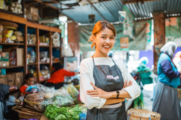 asian female greengrocer standing in front of vegetable stall smiling with crossed arms - obrazy, fototapety, plakaty