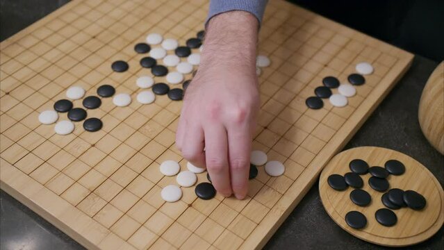 Overhead 45 degree view of gameplay in the ancient Chinese board game Go with a man's hand slowly placing a white stone piece and lingering briefly before committing