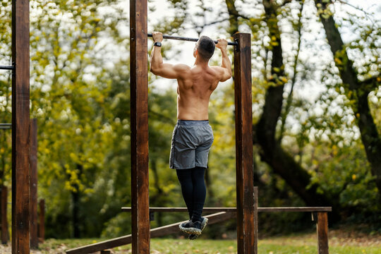 A shirtless muscular bodybuilder is doing pull ups in nature in outdoor gym.