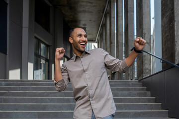 Joyful young man celebrating success with a fist pump gesture on urban stairs. Euphoria,...