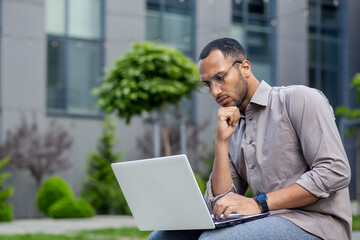 Focused man with glasses working on laptop outdoors, showing concentration and determination in a...