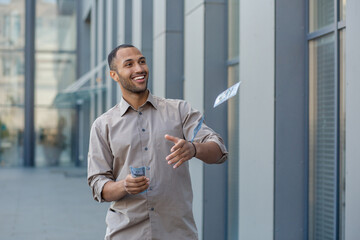 Smiling man casually dressed in beige shirt outside, extending a handshake, holding phone and money, conveying trust and friendliness.