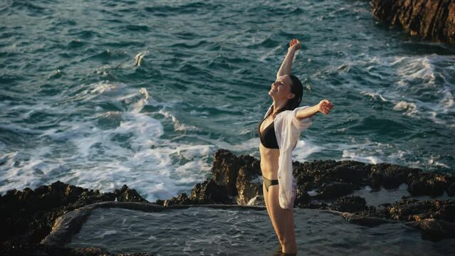A young carefree Caucasian woman splashes water while standing in the natural rock pool with a beautiful sea sunset view.
