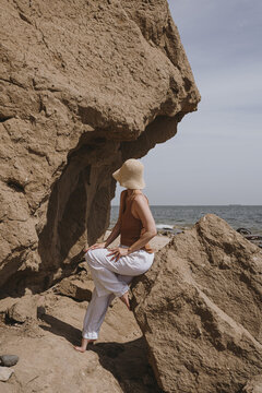Woman in white pants and straw hat sitting on the rock and looking at sea. Elegant fashion, holidays, travel, vacation composition