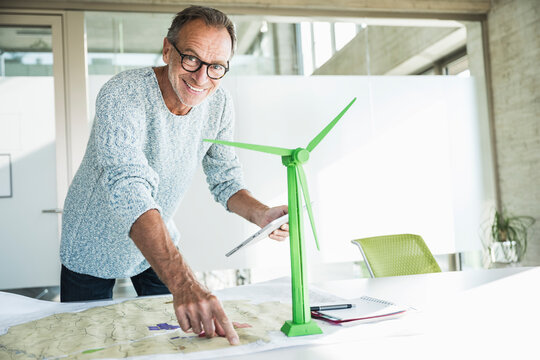 Smiling businessman pointing on blueprint and holding tablet PC near wind turbine model