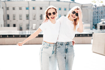 Two young beautiful smiling hipster female in trendy summer white t-shirt and jeans clothes. Carefree women posing in the street. Positive models having fun outdoors. Cheerful and happy. In sunglasses