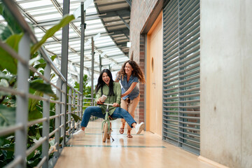 Happy businesswoman enjoys riding bicycle with colleague pushing her in office corridor