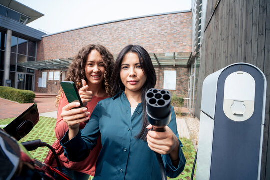 Woman with smart phone and electric plug by fiend at charging station