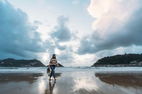Young tourist walking on Playa de la Concha shore at sunset under cloudy sky