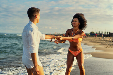 A caucasian man and an african-american woman joyfully dance on the sandy beach, showcasing freedom and happiness.