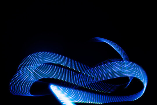 Blue neon curved wave of light as curls, swirl or spiral with dotted stripes on black background, pattern. Abstract background with motion light effect, light painting in holiday style.