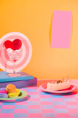 Background with pastel colors. Marshmallow inside a cocktail glass, macarons and cakes on a plate. A piece of paper is stuck on the orange wall. Empty space for text design.