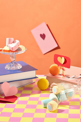 Marshmallow is poured onto the floor from a glass, sweets on a glass pedestal. Scene with pastel colors. Orange background with pink and yellow checkered surface.