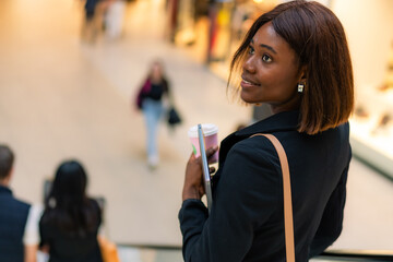 With a laptop, coffee, and handbag, a fashionable young African American woman rides an escalator in the shopping center.