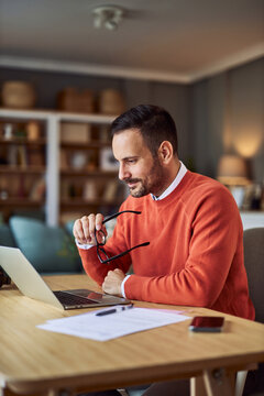 A focused adult man holding his glasses and researching on a laptop at home.